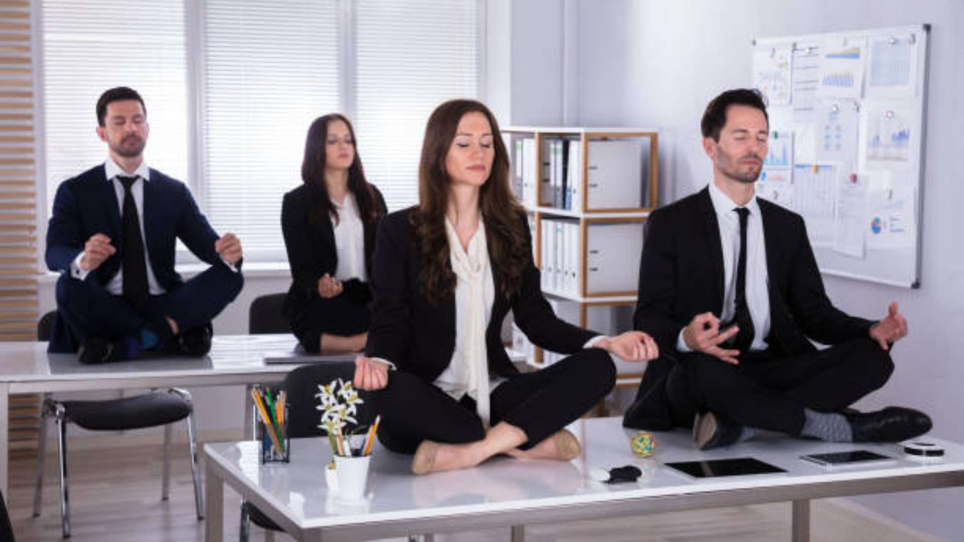 A person in work clothes doing a brief yoga pose beside a desk, bathed in natural light
